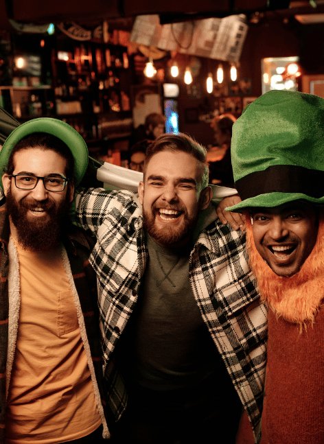 Three bearded men in green leprechaun hats laugh arm-in-arm at a cosy Irish pub bar, capturing St. Patricks Day festive spirit for Dublin hostel party vibes.