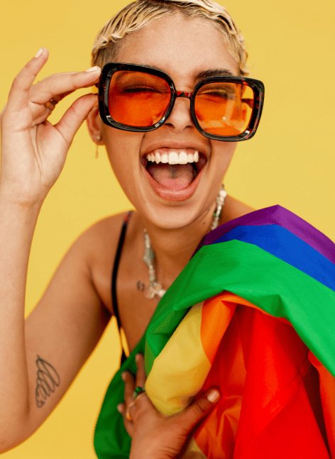 Joyful short-haired woman with bold orange sunglasses laughs exuberantly on yellow background, draped in rainbow Pride flag—vibrant celebration of LGBTQ+ joy.