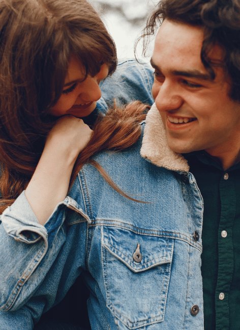 Romantic couple cuddles affectionately outdoors: woman with long brown hair nuzzles bearded man in denim jacket and green shirt against snowy winter backdrop — ideal Valentines vibe.