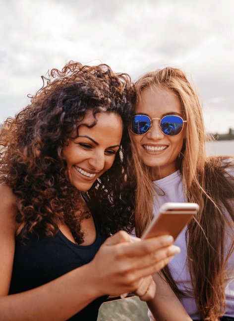 Two smiling diverse women with sunglasses and long hair check smartphone together outdoors by water on sunny day, embodying carefree friendship and adventure.