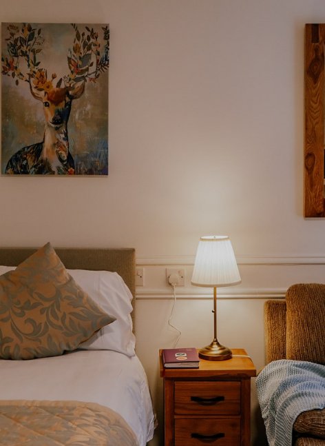 Cosy bedroom at The Maltings Suites with a bed, bedside lamp, armchair and a deer painting on the wall.