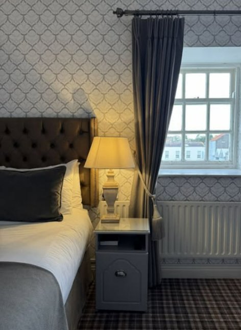 Classic Doolys Hotel bedroom with a tufted headboard, patterned wallpaper, bedside lamp, window with curtains and a grey check carpet.
