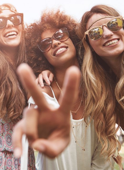 Four friends in Christmas reindeer antlers and festive sweaters laugh around wooden table with drinks at warmly lit holiday pub gathering, red nose on central bearded man.
