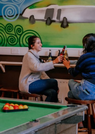 Two friends cheering beers in a living room with a snooker/pool and chatting