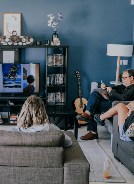 Cosy living room scene: long-haired woman on grey sofa watches tropical beach movie on TV, while glasses-wearing man relaxes nearby with phone amid bookshelves, guitar, and blue decor.