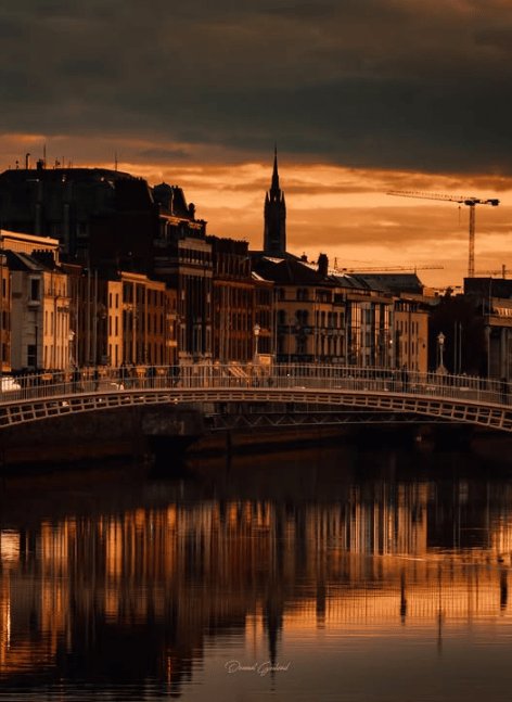Dramatic sunset over Hapenny Bridge in Dublin, with golden spire of Christ Church Cathedral silhouetted against orange sky, reflections shimmering on River Liffey amid historic buildings.