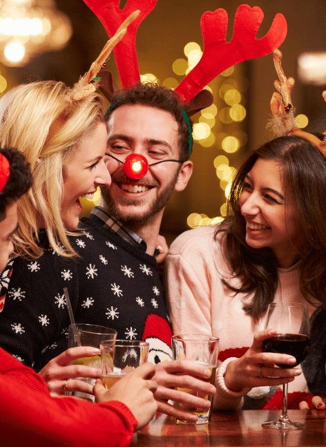 Four friends in Christmas reindeer antlers and festive sweaters laugh around wooden table with drinks at warmly lit holiday pub gathering, red nose on central bearded man.