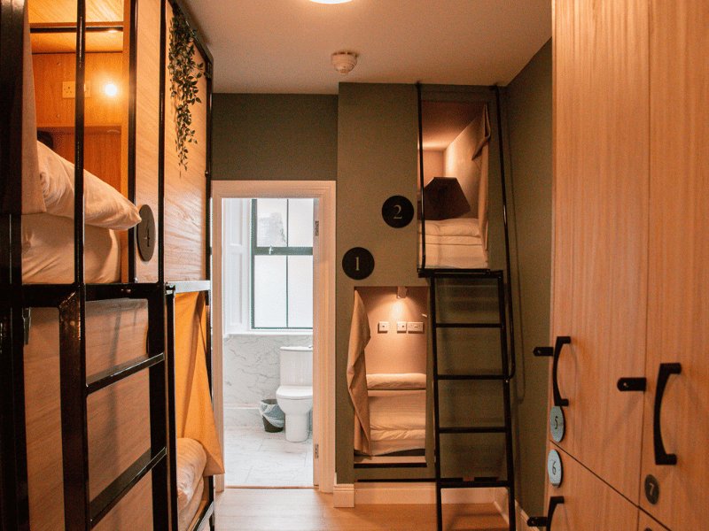Cosy modern hostel bunk room with green walls, wooden bunk beds with yellow blankets, hanging plants, wall lamps and visible ensuite bathroom door