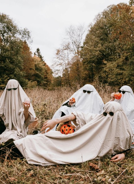 Five friends in white ghost sheets with sunglasses pose playfully in autumn field, holding pumpkins amid orange foliage and tall grasses under cloudy skies.