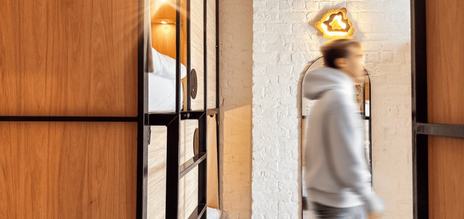 Blurry motion of a guest in hoodie walking past wooden bunk beds with privacy curtains in a bright Gardiner House Hostel dorm room, yellow neon leaf wall lamp glowing on white brick wall.