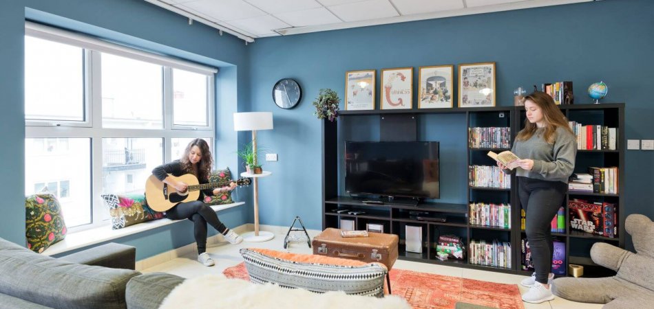 Two girls in a cosy living room, one playing guitar, the other one reading beside the book shelf. 